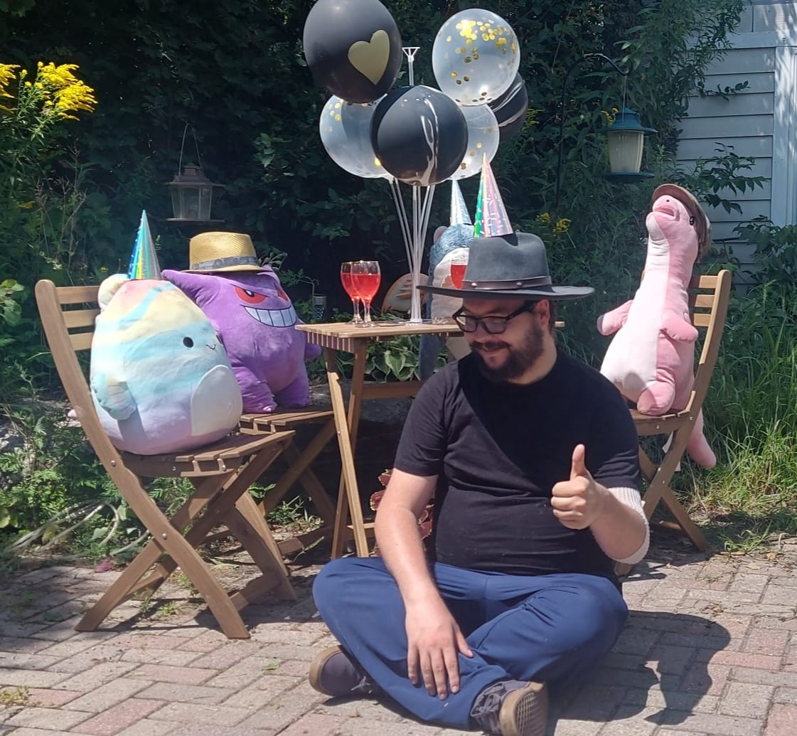 A man with facial hair and a felt sun hat sits on the ground in front of a patio set hosting a stuffed animal patio party, Photo 1